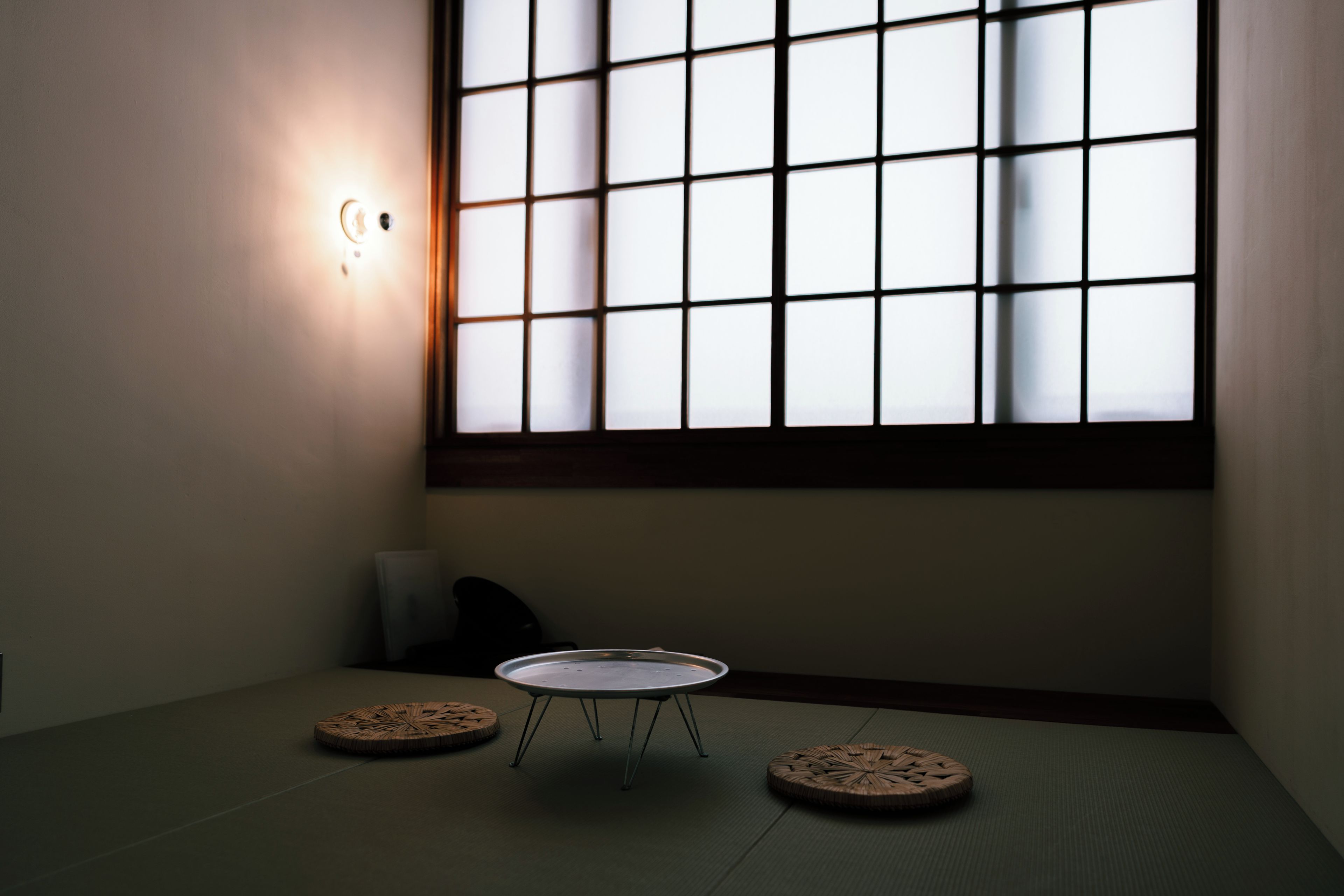 Une pièce avec un sol recouvert de tatamis, une table basse, deux coussins et une grande fenêtre à carreaux.