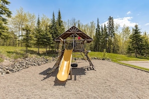 A playground with a slide and climbing structure, surrounded by trees and gravel.