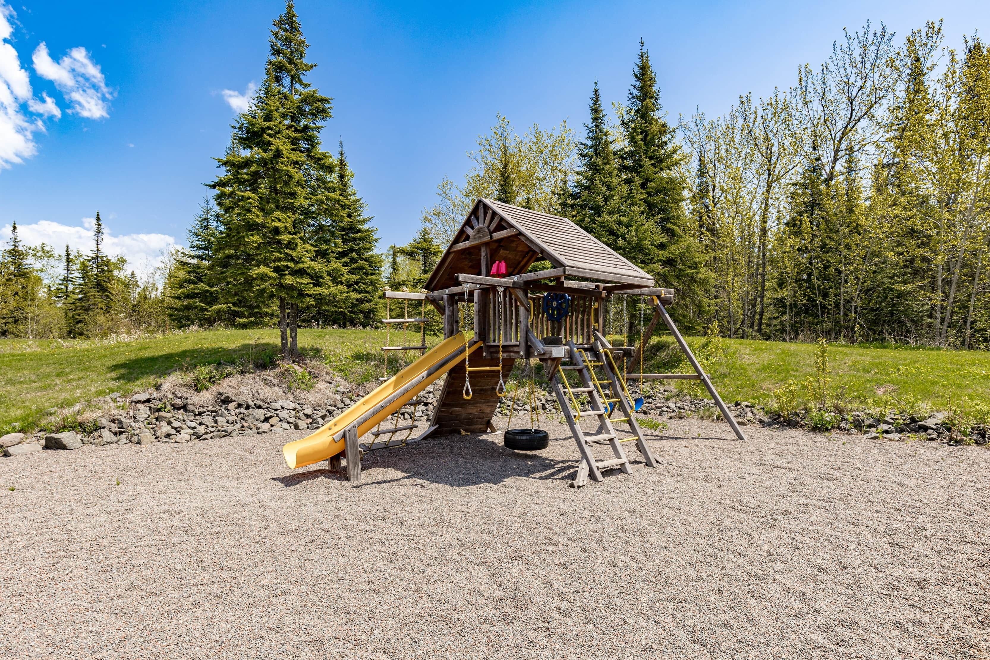 Une structure de jeu en bois, dotée d’un toboggan et d’éléments d’escalade, entourée d’arbres et de gravier.
