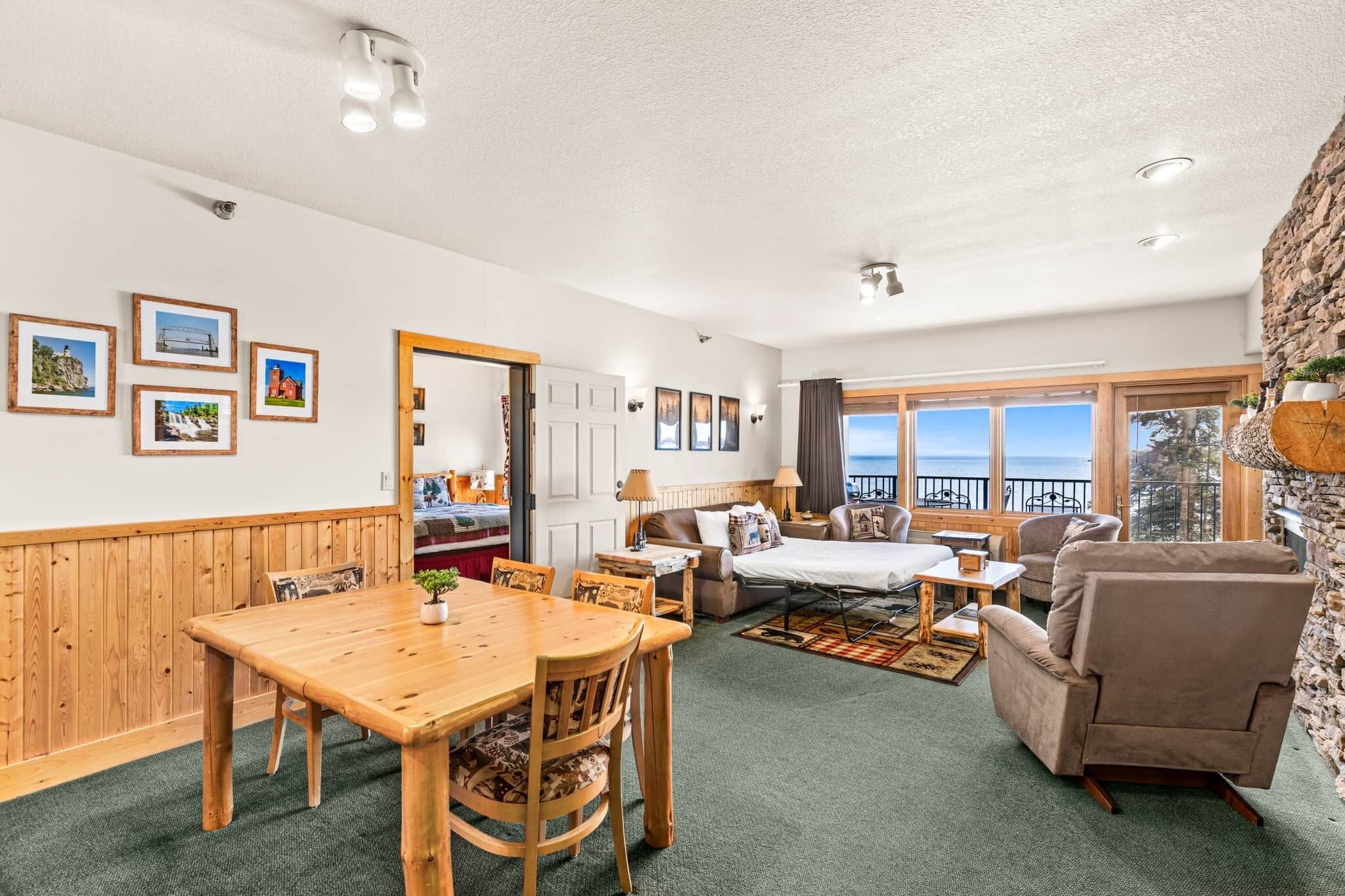 A spacious room with a wooden dining table, a sofa, a chair, and a view of the sea through large windows.