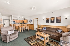 A cozy living room with a brown leather sofa, a wooden coffee table, and a dining area with wooden chairs and a kitchen in the background. - Panoramic Lake Views | 2BR w/King Suite | Pools (Two Harbors)