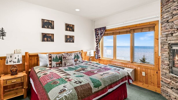 A bedroom with a large bed, a stone fireplace, a window with a view of the sea, and a quilt with animal prints.