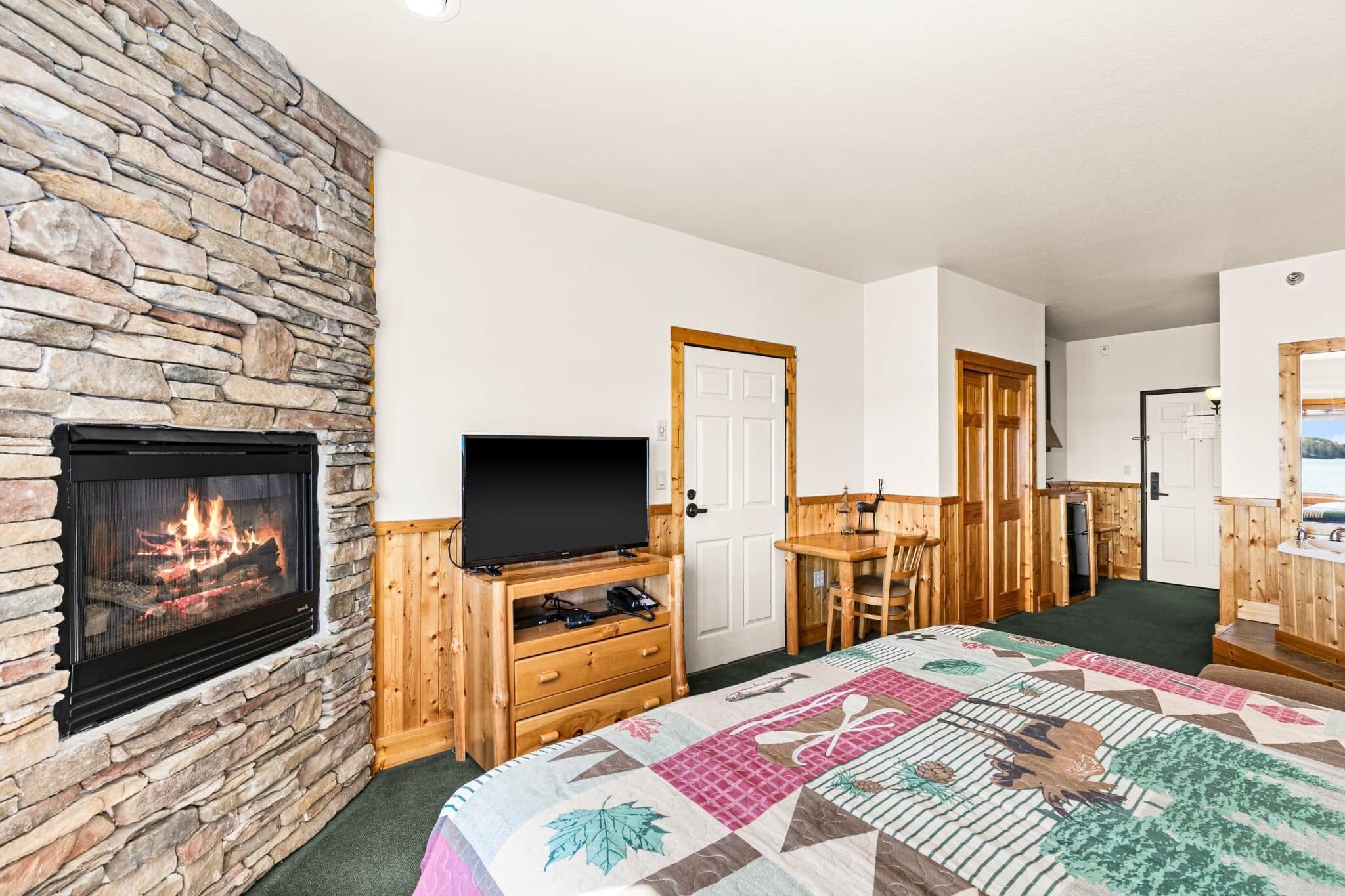 A room with a stone fireplace, a television on a wooden stand, a bed with a quilt, a wooden desk, and a door leading to another room.