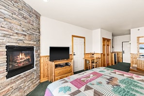 Una habitación con chimenea de piedra, un televisor sobre un mueble de madera, una cama con colcha, un escritorio de madera y una puerta que da a otra habitación.