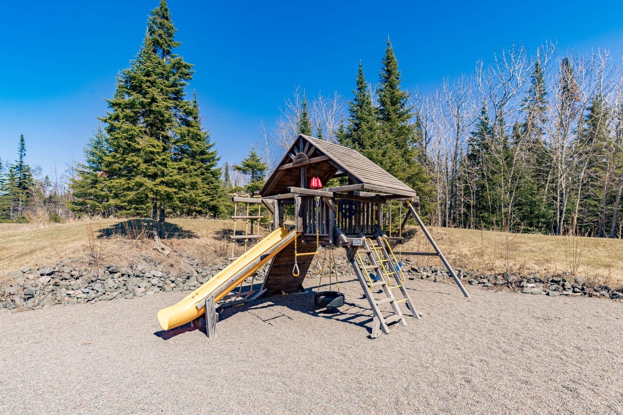 A wooden play structure with a slide and climbing area, surrounded by trees.