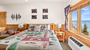 A bedroom with a large bed, a wooden headboard, a view of the sea, and a forest-themed quilt.