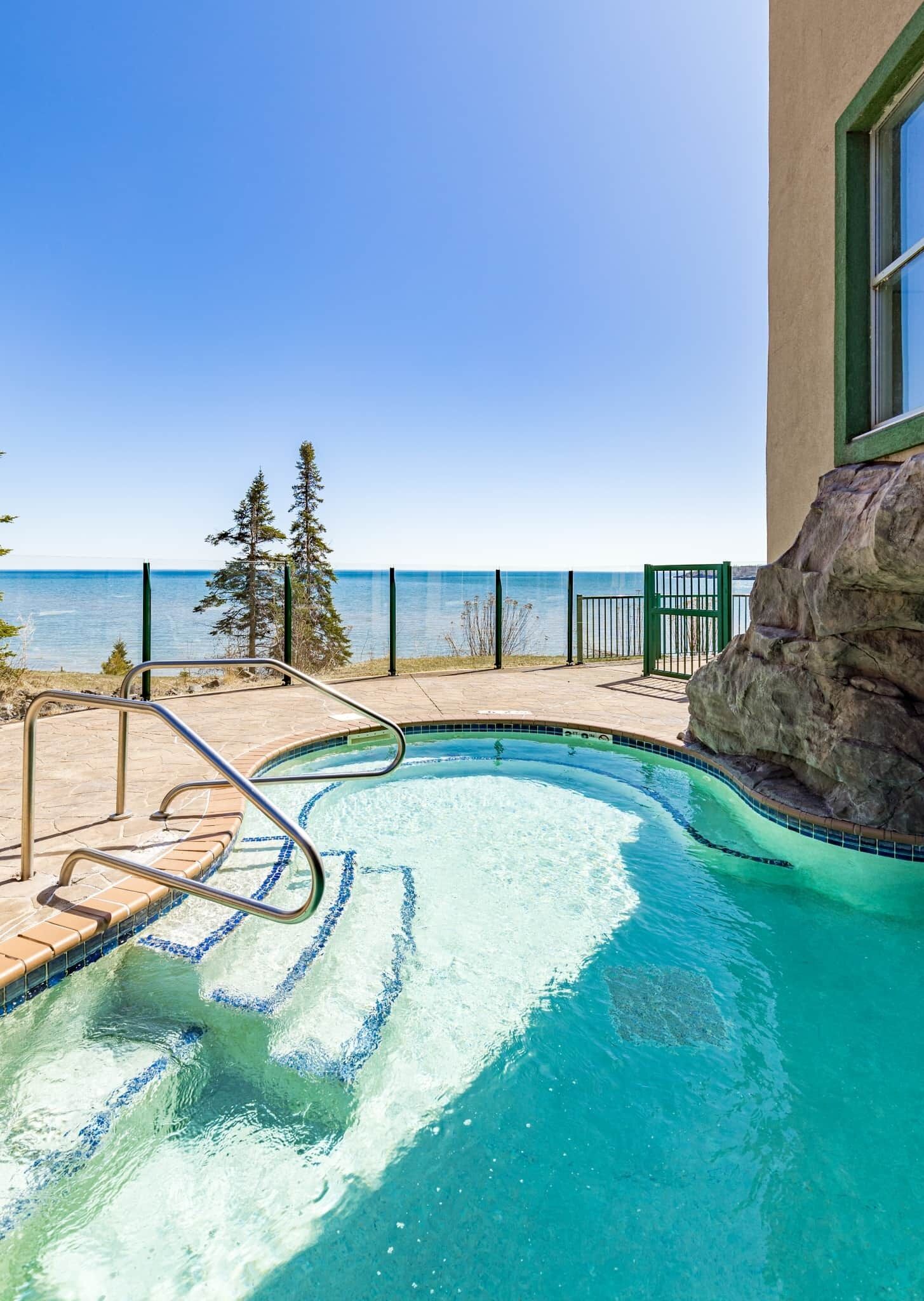 A swimming pool with a view of the sea, surrounded by a stone deck and metal railings.