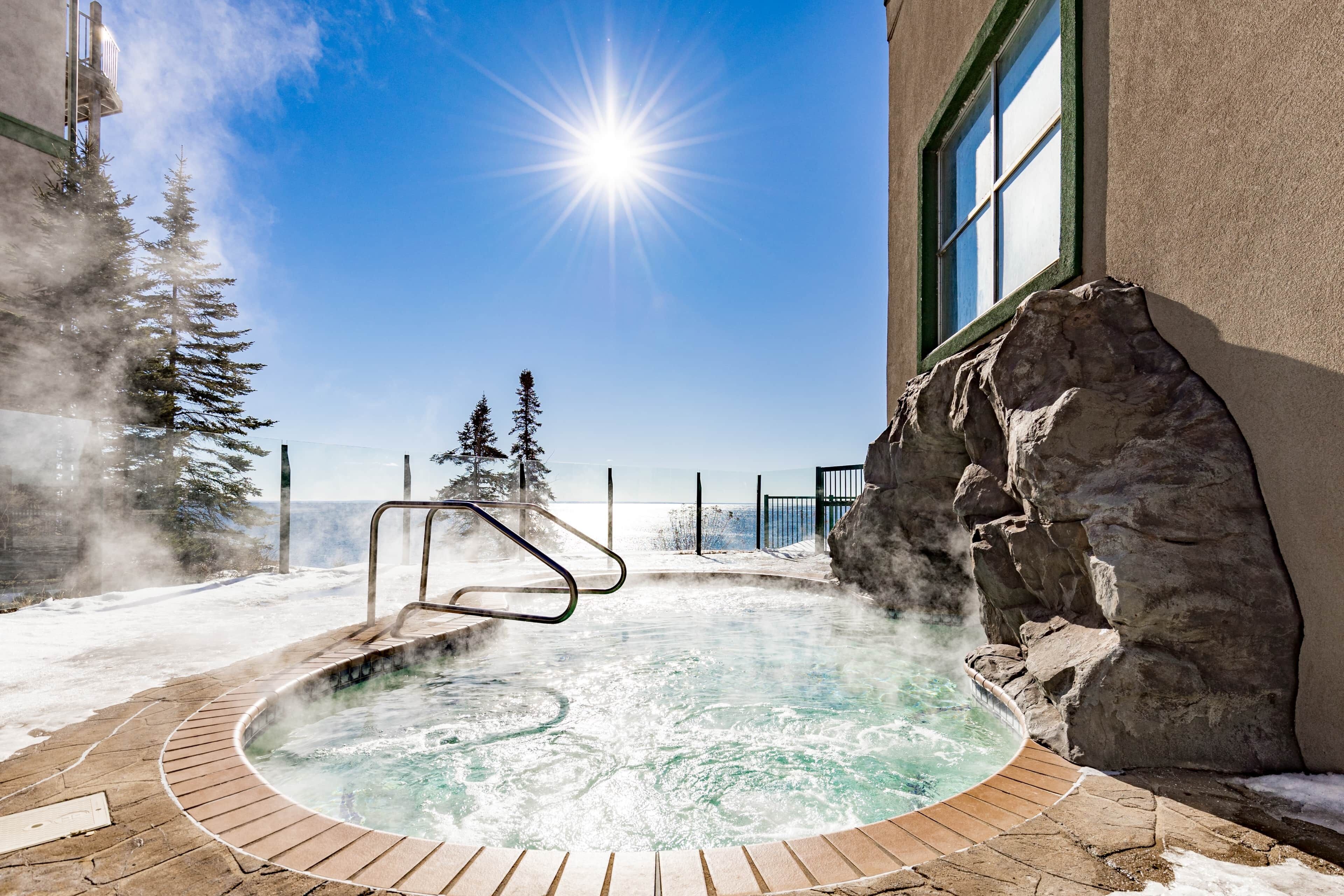 A hot tub with a wooden deck, surrounded by snow and trees, with a clear sky and sun.