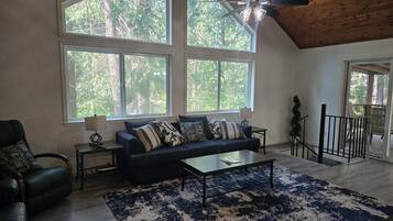 Un salon spacieux avec un plafond haut, de grandes fenêtres, un ventilateur de plafond, un canapé de couleur foncée, une table basse et un tapis à motifs.