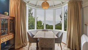 A dining area with a table and chairs, a wooden cabinet, and a view through large windows. - Waitati Retreat - Coastal Calm Overlooking Blueskin Bay (Waitati)
