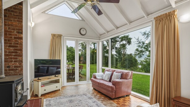 A spacious living room with a fireplace, a television, a sofa, and large windows offering a view of the outdoors. - Waitati Retreat - Coastal Calm Overlooking Blueskin Bay (Waitati)