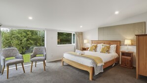 A bedroom with a bed, two chairs, a nightstand, a wooden wardrobe, and a window with a view of greenery. - Waitati Retreat - Coastal Calm Overlooking Blueskin Bay (Waitati)