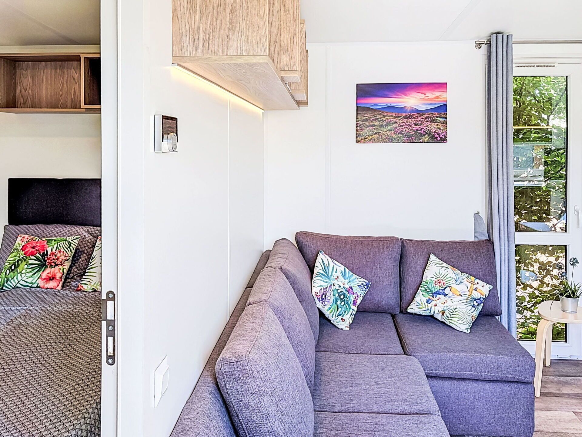 Un salon moderne avec un canapé gris, des coussins à motifs floraux, un tableau accroché au mur et une porte-fenêtre coulissante donnant sur un balcon.