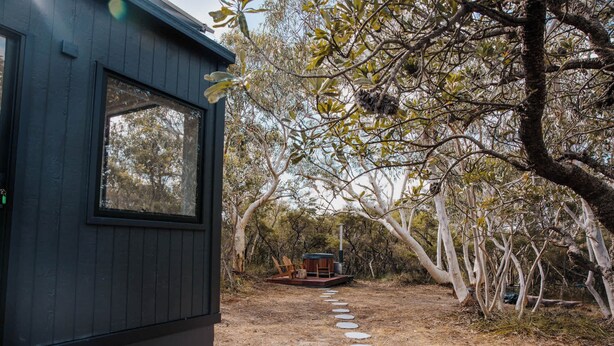 Una piccola baita con una veranda, immersa tra gli alberi e un sentiero sterrato che conduce a una panchina.