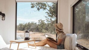 Une personne est assise sur un lit, en train de lire un livre. On aperçoit, par la fenêtre, des arbres, et une petite table porte une lanterne.