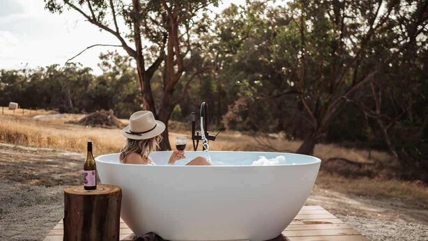 Une personne se détend dans une grande baignoire blanche à l’extérieur, avec une bouteille de vin et un chapeau posé sur un tabouret en bois à proximité.