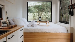 Ein Schlafzimmer mit einem Bett, einem Nachttisch aus Holz, einem Fenster mit Blick auf Bäume und einem an der Wand hängenden Kleiderhaken.