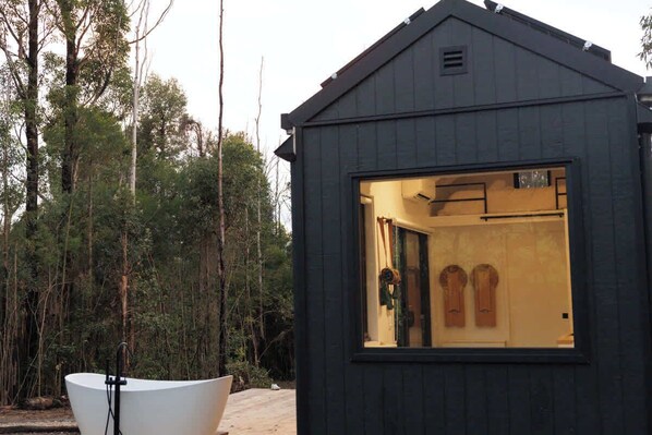 Une petite cabane en bois sombre, dotée d’une grande fenêtre qui donne sur un intérieur comprenant une porte et deux panneaux en bois.