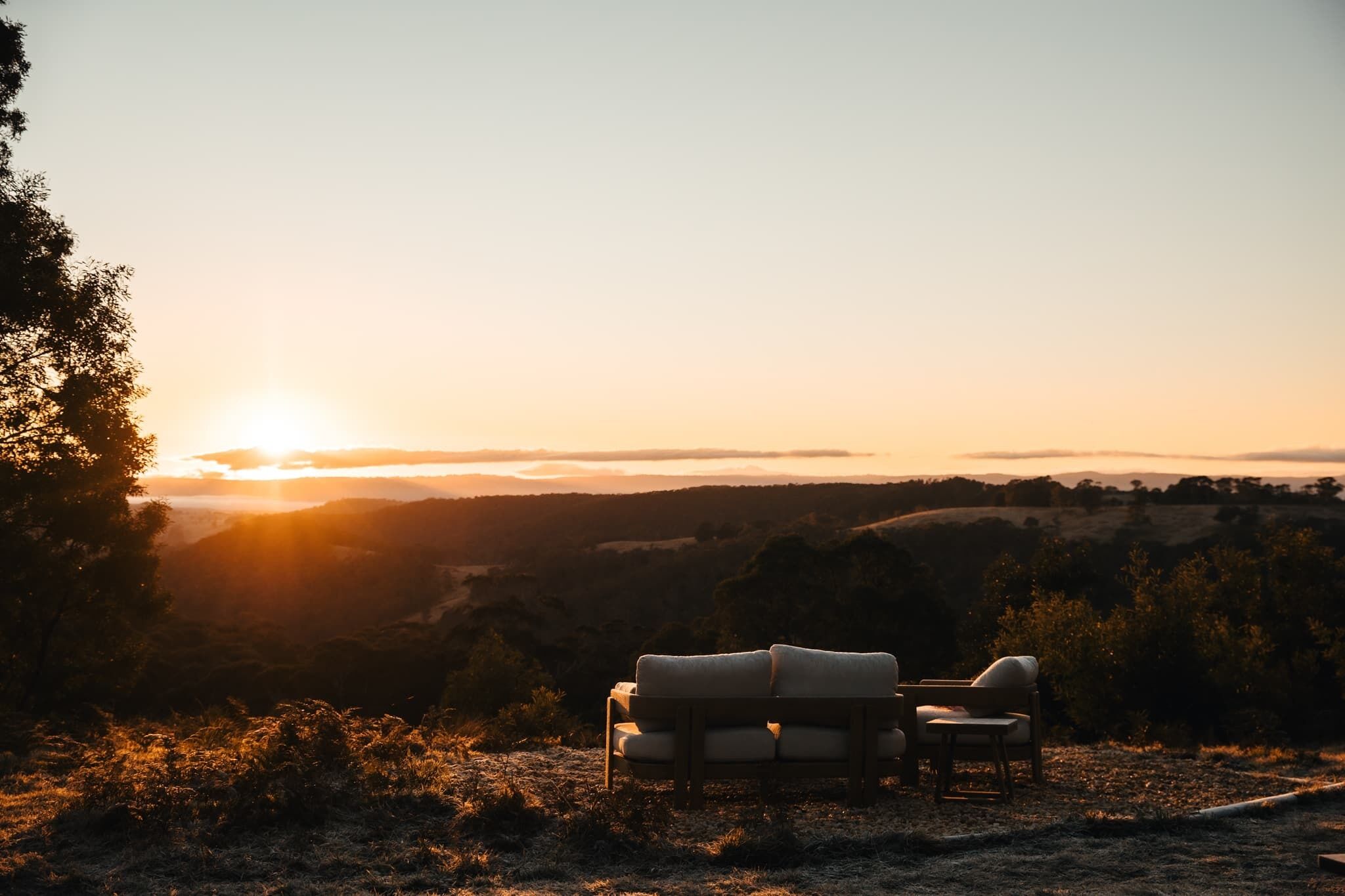 A sunset view with a sofa and chair on a hill overlooking a valley.