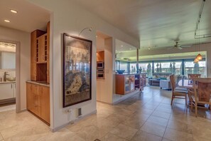 A modern kitchen with wooden cabinets, a large framed artwork, and a dining area with a table and chairs. - Kapalua Ridge | Ocean View 5 BR, Sleeps 14 | Car Incl. w/ 6+ Nights | KRV ML-3948 by KBM (Lahaina)