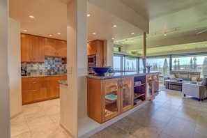 A modern kitchen with wooden cabinets and a large island, a dining area with a sofa and table, and a view of trees through large windows. - Kapalua Ridge | Ocean View 5 BR, Sleeps 14 | Car Incl. w/ 6+ Nights | KRV ML-3948 by KBM (Lahaina)