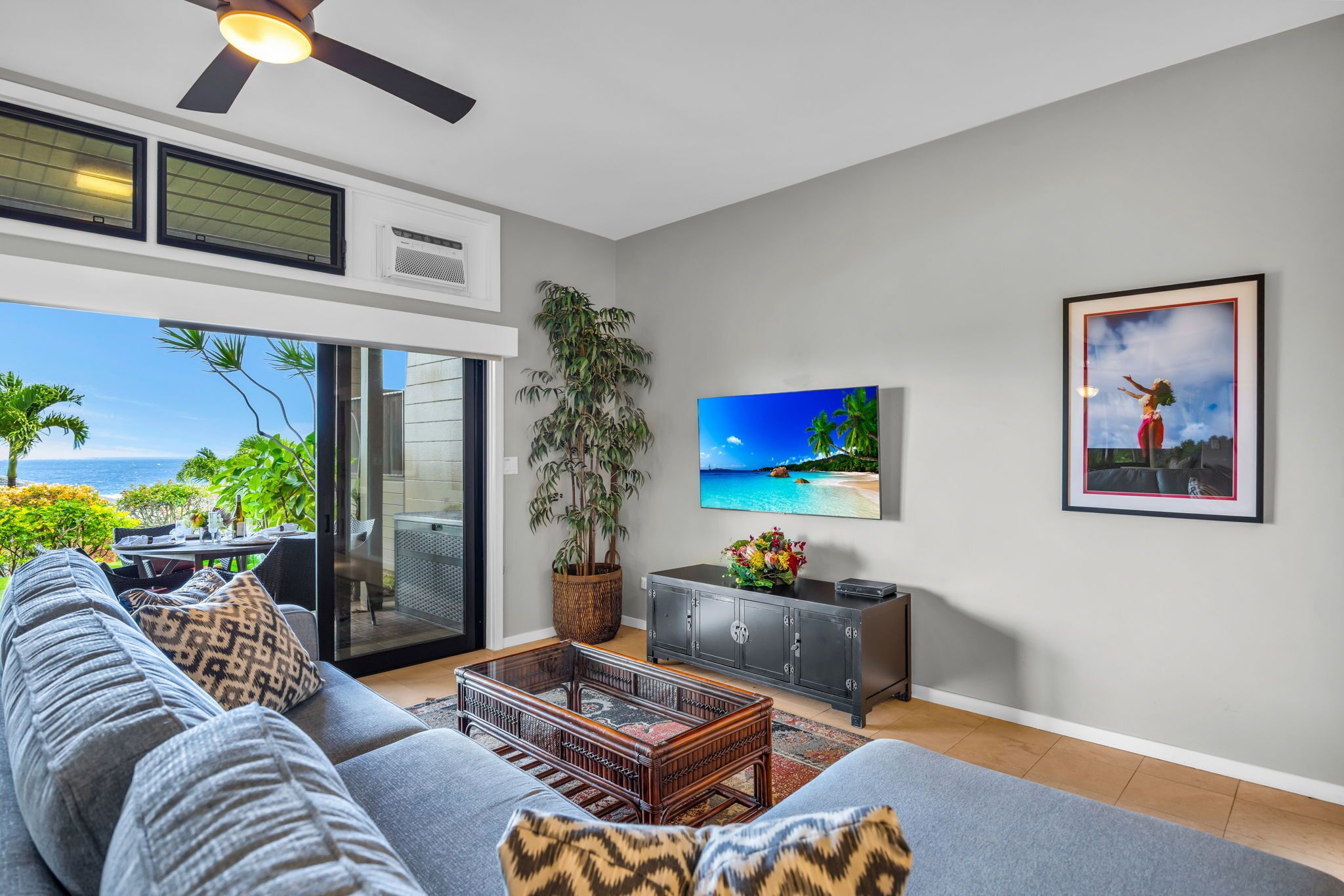 A modern living room with a large gray sofa, a coffee table, and a television. There is a large window with a view of the ocean and palm trees.