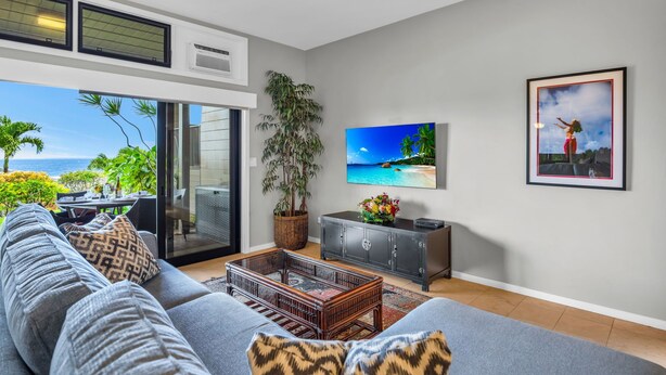 Ein modernes Wohnzimmer mit einem großen grauen Sofa, einem Couchtisch und einem Fernseher. Ein großes Fenster bietet Blick auf den Ozean und Palmen.