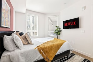 A bedroom with a bed, pillows, a blanket, a TV, and a plant.