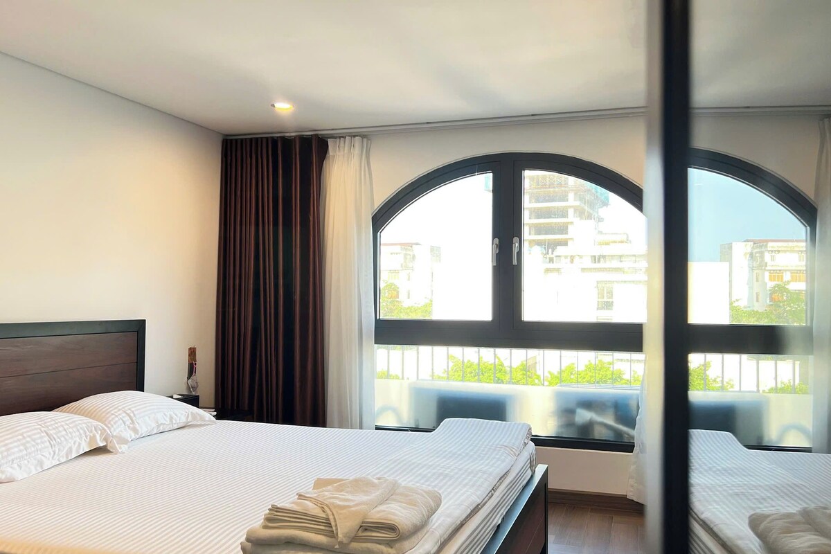 Ein Hotelzimmer mit einem großen Bogenfenster, einem Bett mit weißer Bettwäsche und einem Nachttisch.
