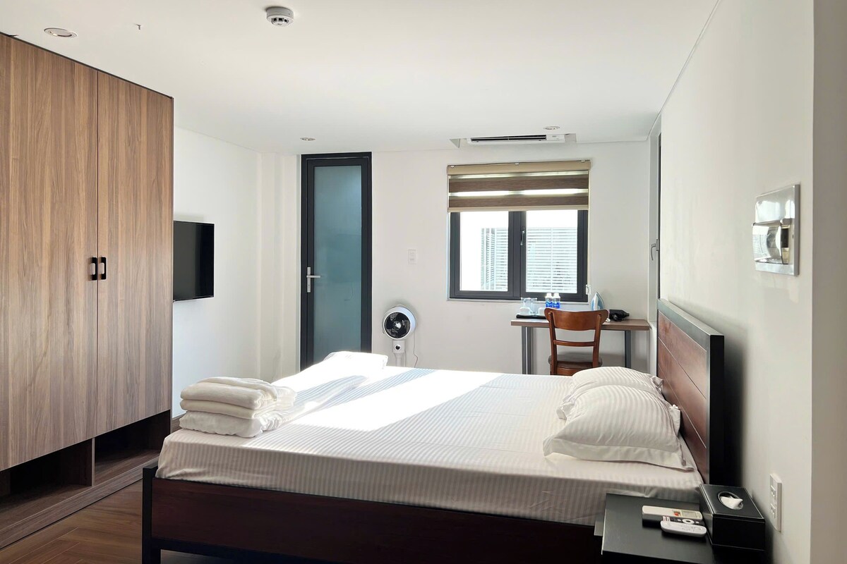 Ein Hotelzimmer mit Bett, Schreibtisch, Stuhl, Kleiderschrank, Fernseher, einem Fenster mit Jalousien und einer Decken-Klimaanlage.