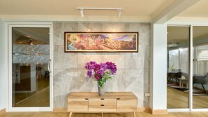 A modern living room with a marble wall, a wooden console table, a framed artwork, and a vase of purple flowers. - TEN-Place Srinakarin (Bangkok)