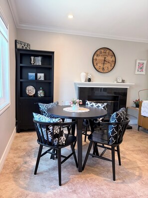 A dining area with a round table, black chairs with patterned cushions, a black bookshelf, a wall clock, and a fireplace.