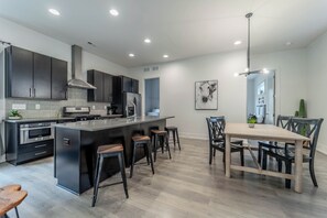 A modern kitchen with dark cabinetry, a central island, bar stools, and a dining table with chairs. - Serene 4BR Home with Spacious Backyard (Charlotte)