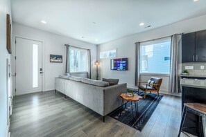 A modern living room with a grey sofa, a small round table with a plant, a fireplace, and a kitchen area with cabinets and a TV. - Serene 4BR Home with Spacious Backyard (Charlotte)