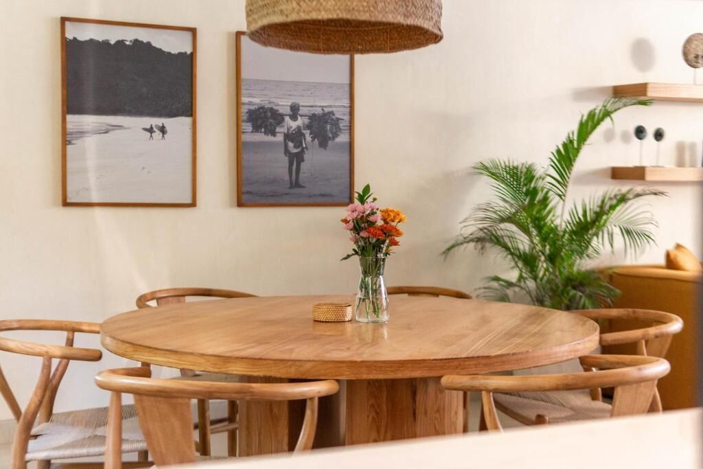 A round wooden dining table with six chairs, a vase with flowers, and two framed photographs on the wall.
