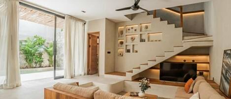 A modern living room with a sofa, a coffee table, and a TV. There is a staircase with a TV on the upper landing.