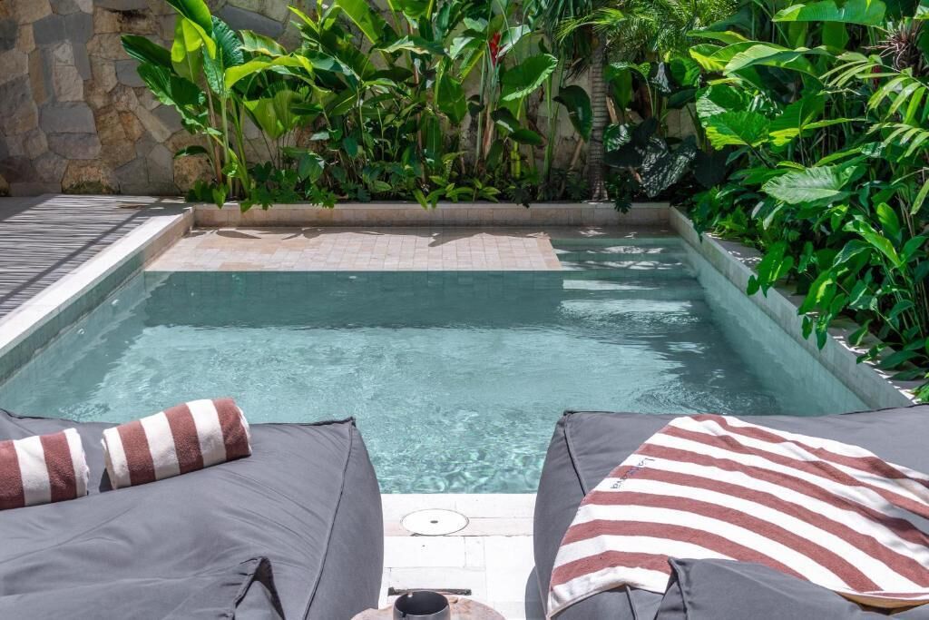 A rectangular pool with clear water, surrounded by lush greenery and a stone wall. There are two lounge chairs with striped cushions.