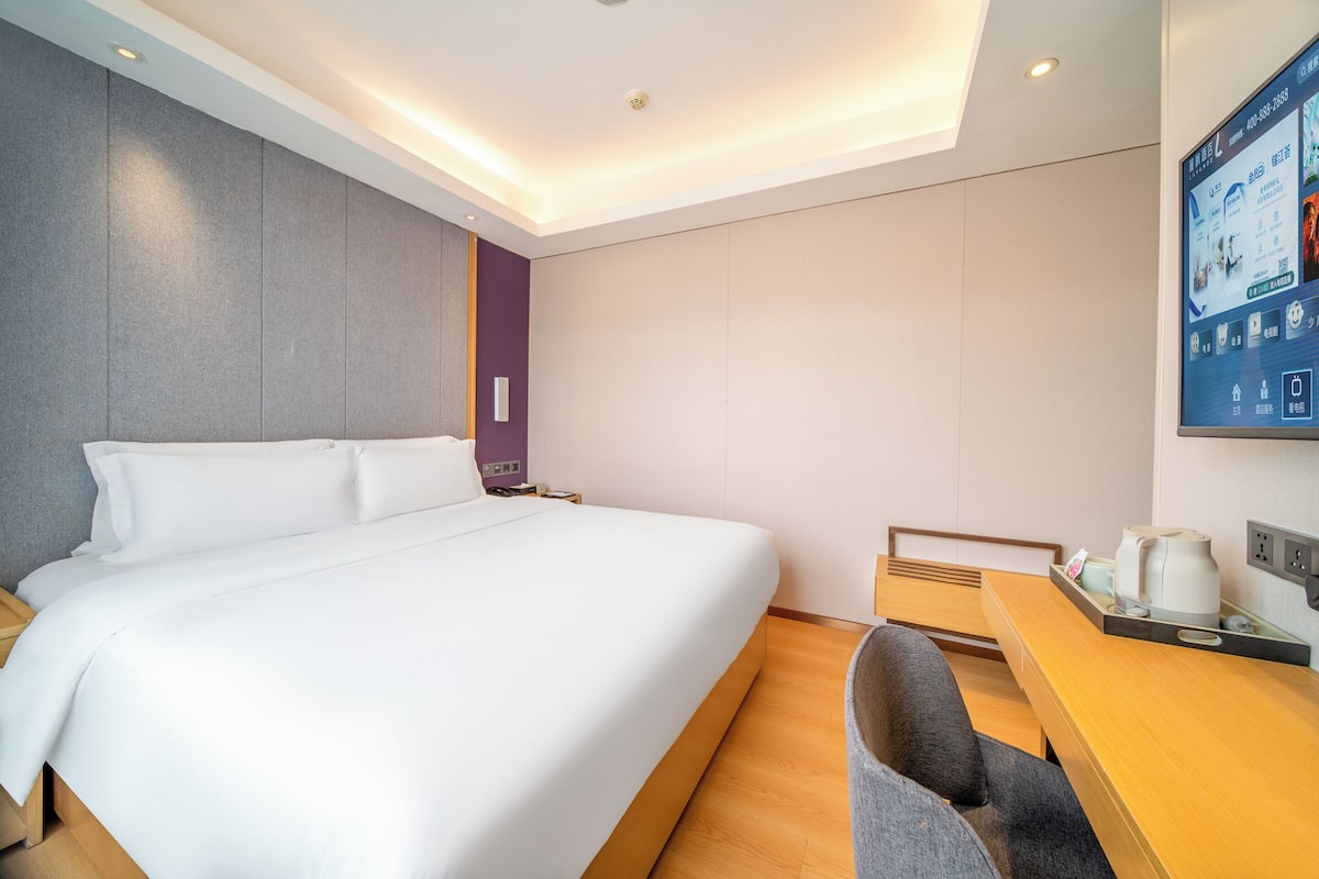 Habitación de hotel con una cama grande, un escritorio con cafetera y una pantalla en la pared mostrando información de la habitación.