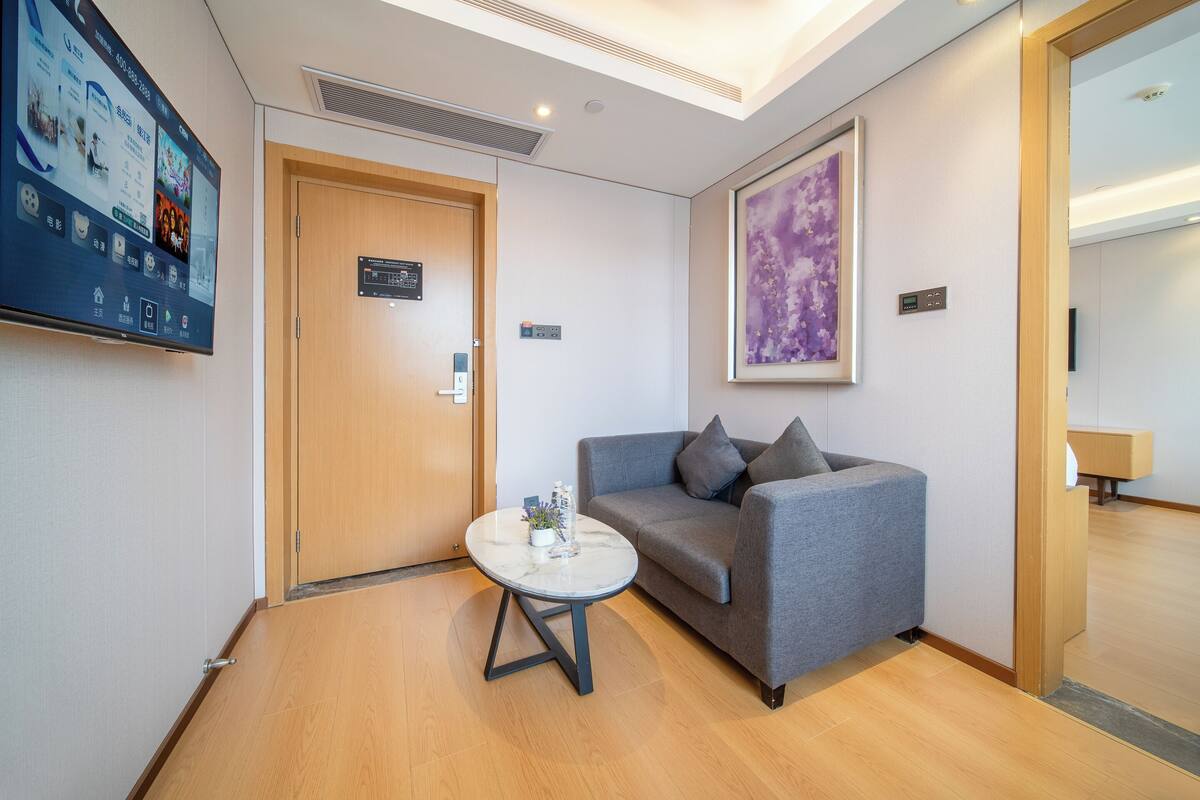 Un moderno vestíbulo de hotel con un sofá gris, una mesa redonda con un florero, un televisor montado en la pared y una puerta de madera.