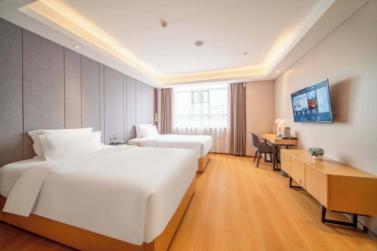 Quarto de hotel com duas camas, uma escrivaninha, uma cadeira, uma televisão e uma janela grande.