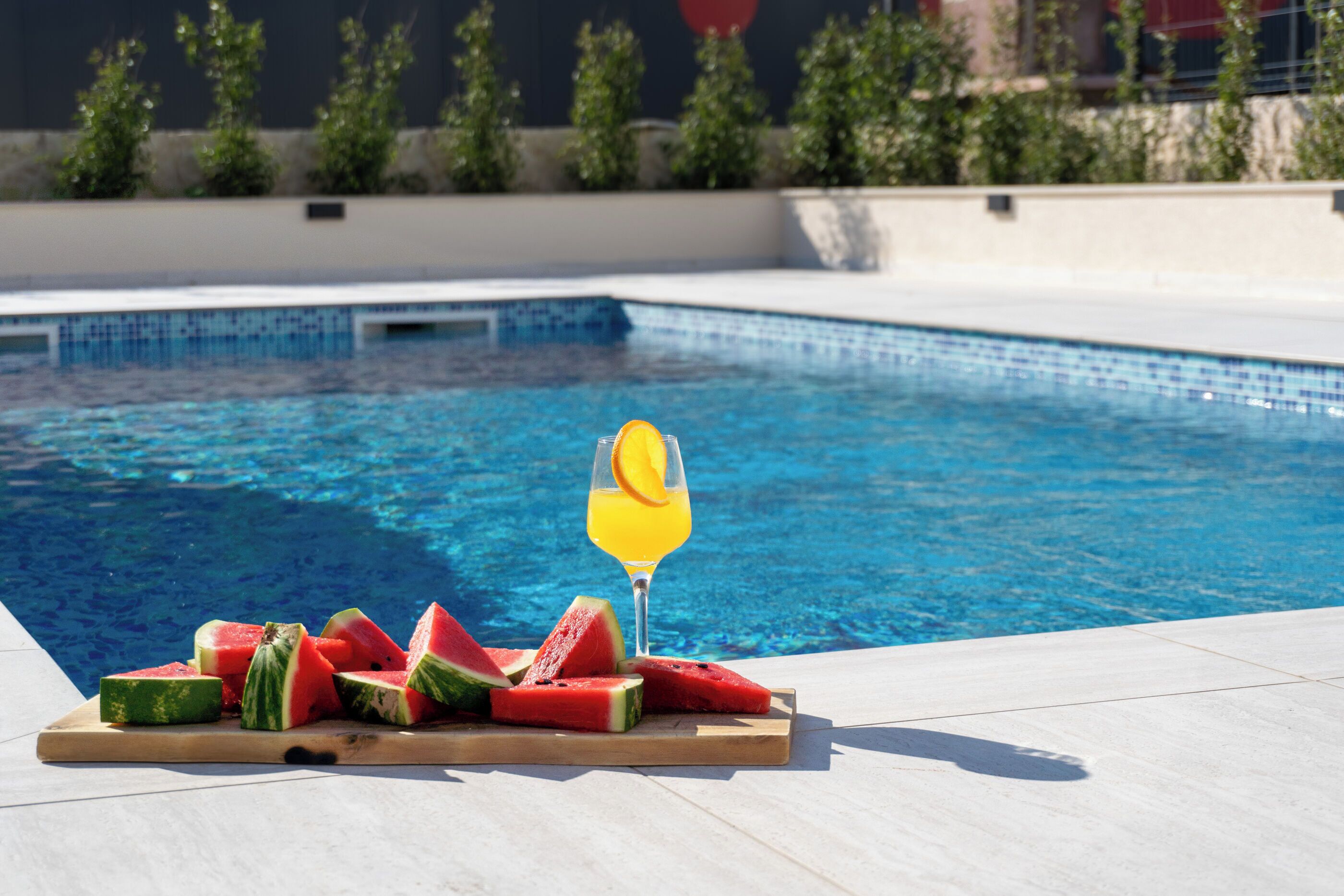 Une piscine avec une planche en bois sur laquelle sont disposées des tranches de pastèque et un verre de jus d’orange.