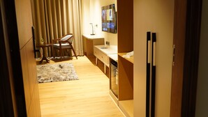 A narrow hallway with wooden flooring, a dining area with a table and chairs, a kitchen area with a refrigerator, and a television mounted on the wall. - Luxe Manor (Kharar)