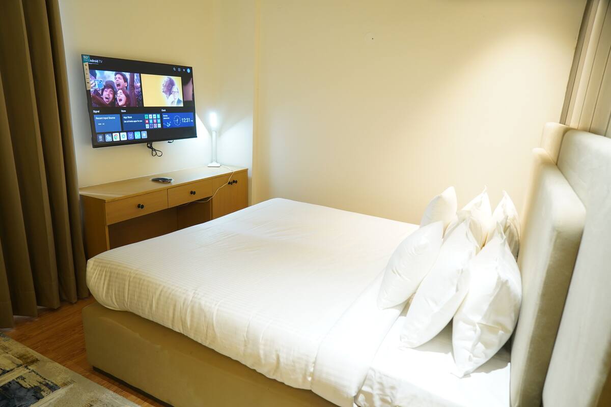 Ein Hotelzimmer mit einem Bett, einem an der Wand befestigten Fernseher, einem Nachttisch mit Lampe und einem Holznachtkästchen.