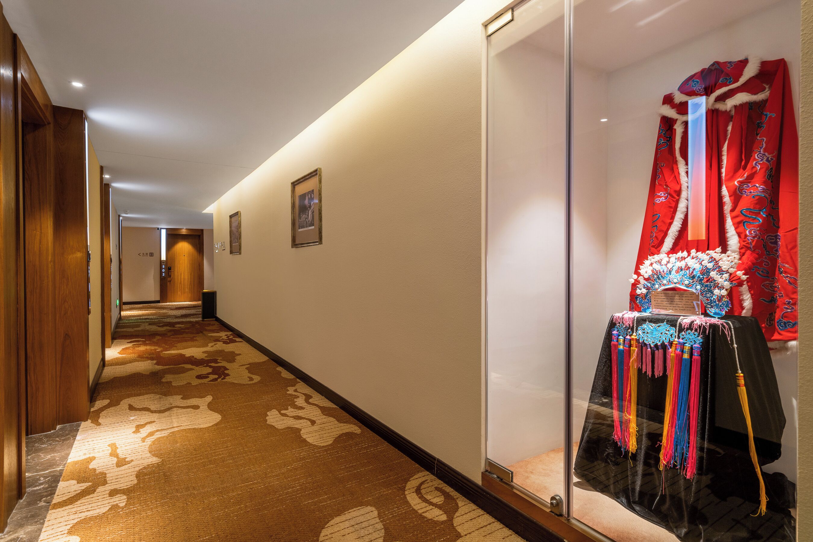 Pasillo de hotel con alfombra estampada, puertas de madera y una vitrina de cristal con cortina roja.