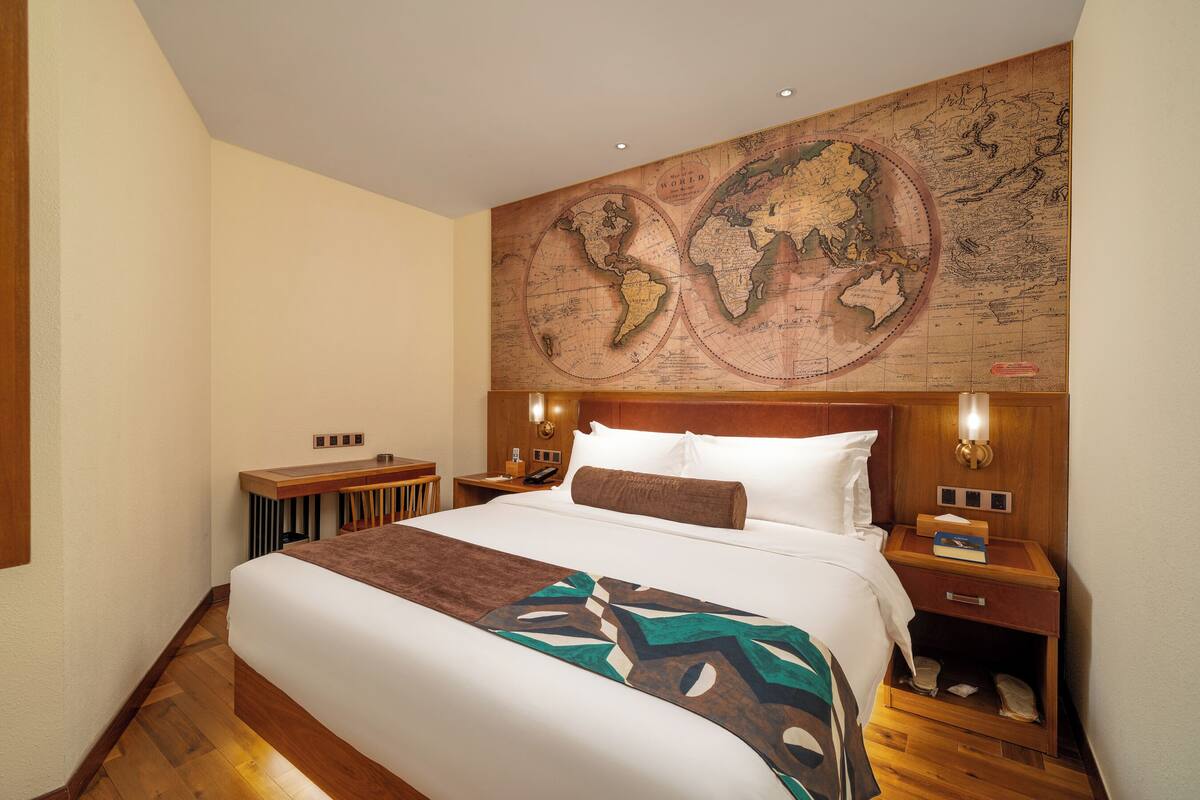 A hotel room with a large bed, wooden headboard, a world map on the wall, a desk, and a nightstand with a lamp.