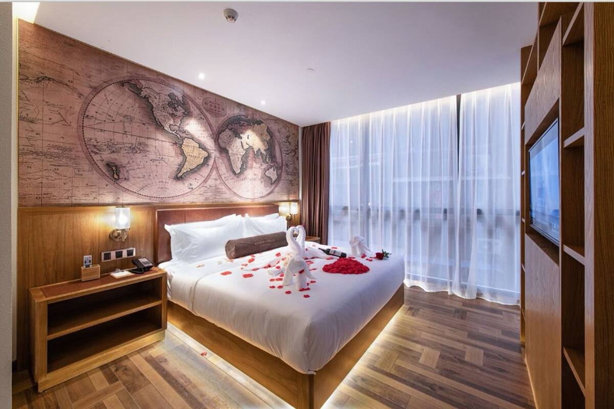 A hotel room with a large bed, wooden headboard, a world map on the wall, a bedside table with a lamp, and a large window with sheer curtains.