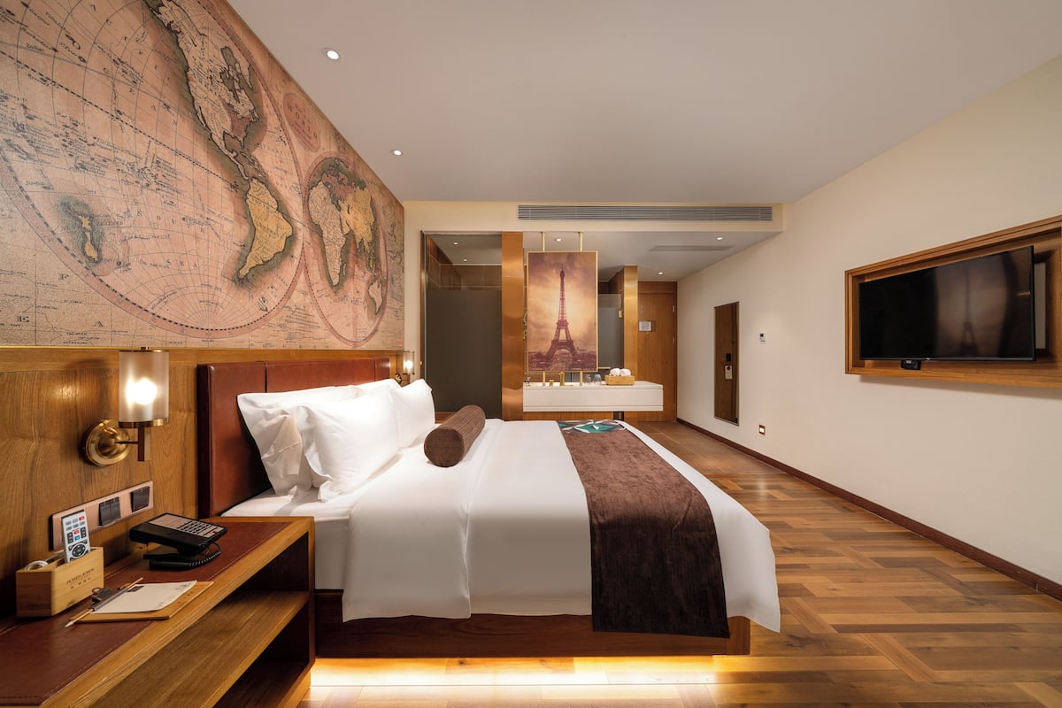 A hotel room with a large map on the wall, a bed with white linens, a wooden nightstand with a phone and notepad, and a TV mounted on the wall.
