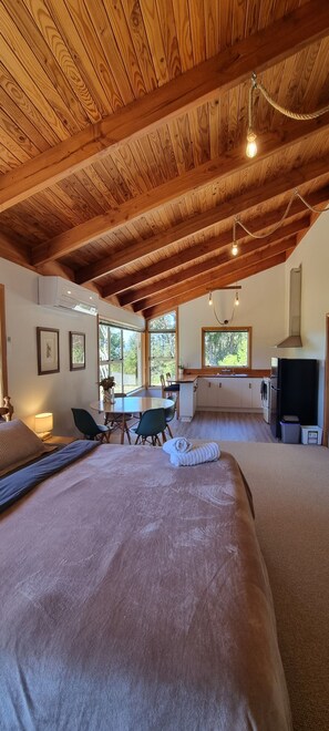 Una habitación con techo de madera, una cama, una mesa de comedor, sillas, un televisor y una ventana con vistas a árboles.