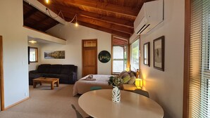 Ein gemütliches Wohnzimmer mit einer Couch, einem Couchtisch und einem Esstisch. Eine Tür führt in einen anderen Raum, ein Gemälde hängt an der Wand und ein Fenster mit Jalousien ist vorhanden.