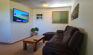 Un salón con un sofá modular marrón, una mesa de centro de madera, un televisor montado en la pared, un cuadro enmarcado y una ventana con persianas.
